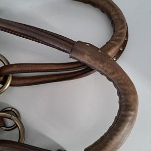May include: A brown leather handbag handle featuring a padded, textured grip section with visible stitching and small metal studs. The handle is attached to the bag body using large, circular, antique-toned brass rings that connect multiple leather straps.