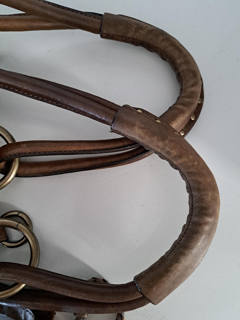 May include: A brown leather handbag handle featuring a padded, textured grip section with visible stitching and small metal studs. The handle is attached to the bag body using large, circular, antique-toned brass rings that connect multiple leather straps.