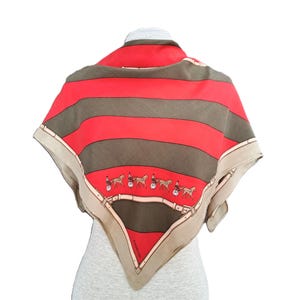 May include: A square silk scarf with a red and brown striped pattern. The scarf features a border with a horse-drawn carriage design and a tan trim. The scarf is draped over a mannequin.