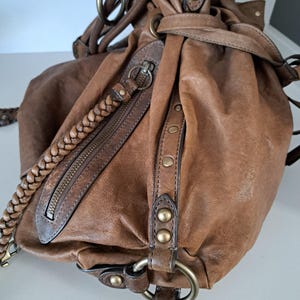 May include: A brown leather hobo bag featuring a braided shoulder strap, a diagonal zipped pocket, and side panels accented with brass-toned metal studs and circular rings. The bag has a gathered, slouchy silhouette with visible stitching details.