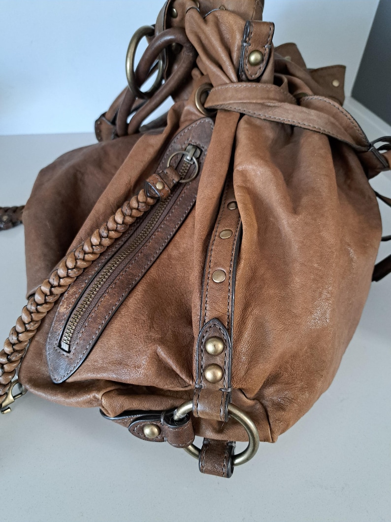 May include: A brown leather hobo bag featuring a braided shoulder strap, a diagonal zipped pocket, and side panels accented with brass-toned metal studs and circular rings. The bag has a gathered, slouchy silhouette with visible stitching details.