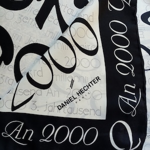 May include: A white and black silk scarf with the text "An 2000" and "Daniel Hechter Paris" printed on it. The scarf has a design of repeating circles and the number 2000.
