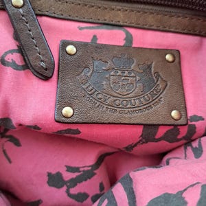 May include: The interior of a handbag featuring a pink fabric lining with a black repeating pattern. A rectangular brown leather patch is attached with four brass-toned rivets, embossed with the text Juicy Couture Born in the Glamorous USA.