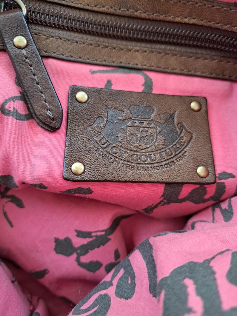 May include: The interior of a handbag featuring a pink fabric lining with a black repeating pattern. A rectangular brown leather patch is attached with four brass-toned rivets, embossed with the text Juicy Couture Born in the Glamorous USA.