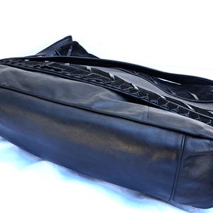 May include: A black leather shoulder bag featuring a smooth leather base and a flap closure with a textured suede panel. The flap is accented with a decorative braided leather trim that runs horizontally across the center of the bag's front surface.