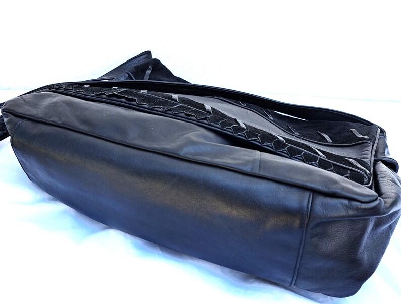 May include: A black leather shoulder bag featuring a smooth leather base and a flap closure with a textured suede panel. The flap is accented with a decorative braided leather trim that runs horizontally across the center of the bag's front surface.