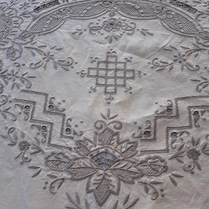 May include: A white tablecloth with intricate embroidered designs in a light brown color. The design features a central floral motif surrounded by geometric patterns and smaller floral details.