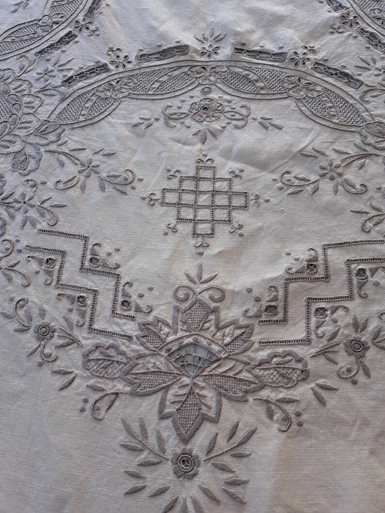 May include: A white tablecloth with intricate embroidered designs in a light brown color. The design features a central floral motif surrounded by geometric patterns and smaller floral details.