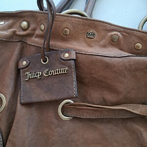 May include: A brown leather handbag featuring a hanging rectangular tag with the text Juicy Couture in gold metal lettering. The bag includes brass-toned metal studs, a small crown emblem, and a leather drawstring threaded through metal grommets.