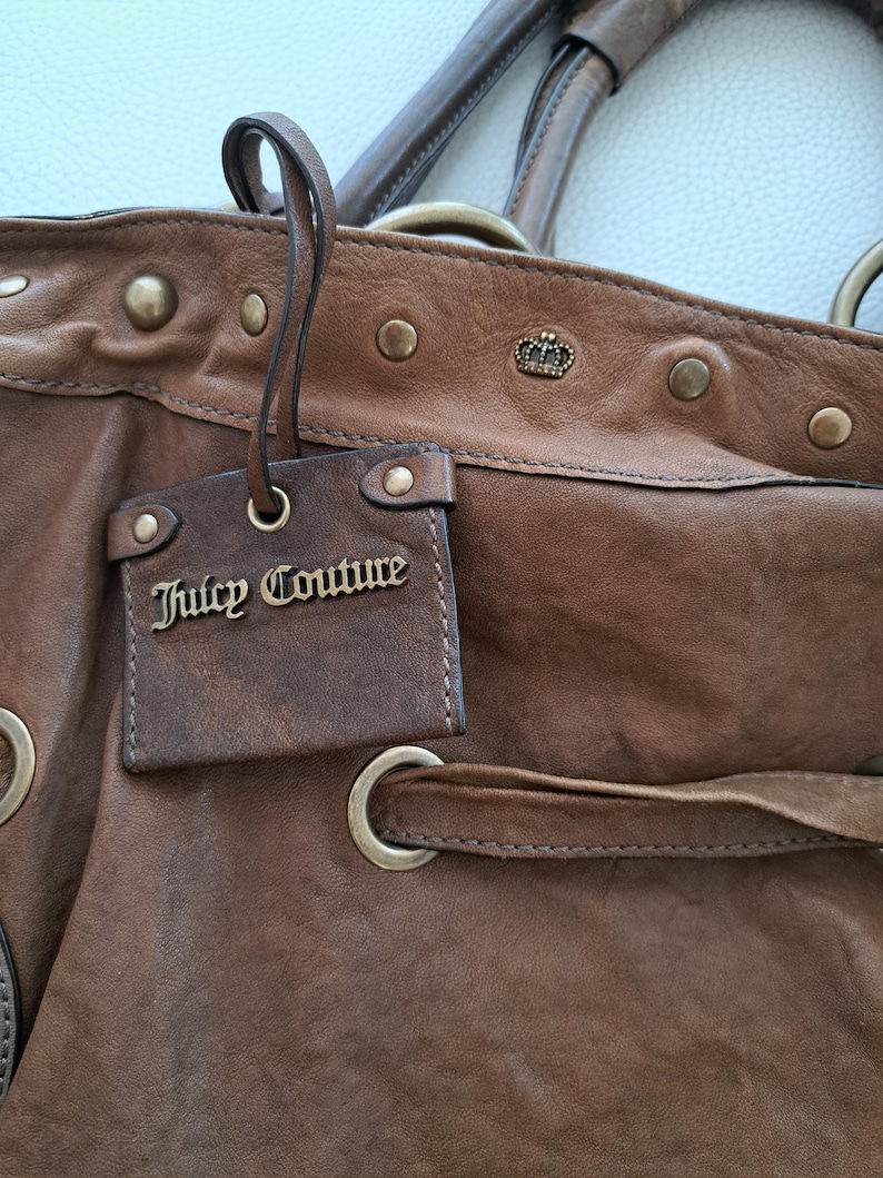 May include: A brown leather handbag featuring a hanging rectangular tag with the text Juicy Couture in gold metal lettering. The bag includes brass-toned metal studs, a small crown emblem, and a leather drawstring threaded through metal grommets.