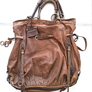 May include: A brown leather hobo bag featuring a drawstring closure, two braided zip pulls, and a rectangular tag embossed with the text Juicy Couture. The bag includes dual top handles, brass-toned hardware, and a gathered, slouchy silhouette.