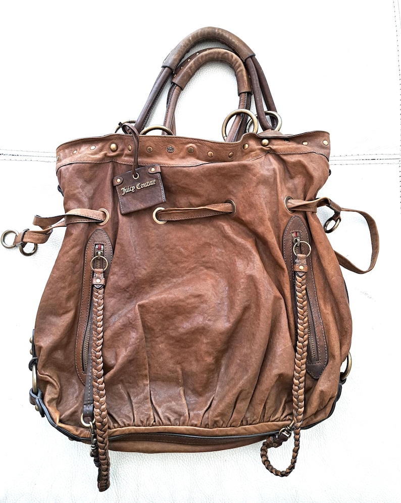 May include: A brown leather hobo bag featuring a drawstring closure, two braided zip pulls, and a rectangular tag embossed with the text Juicy Couture. The bag includes dual top handles, brass-toned hardware, and a gathered, slouchy silhouette.