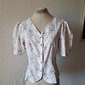 Op de afbeelding: Een wit blouse met een bloemenprint met blauwe en roze bloemen. Het blouse heeft een v-hals en een knoopsluiting aan de voorkant.