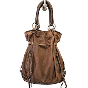 May include: A brown leather hobo bag featuring a drawstring closure, two vertical zipped pockets on the front, and braided leather accents. The bag includes a single top handle attached with large metal rings and additional side hardware details.