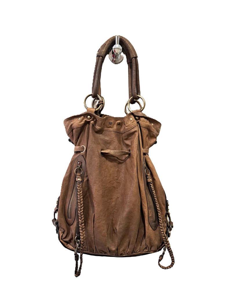May include: A brown leather hobo bag featuring a drawstring closure, two vertical zipped pockets on the front, and braided leather accents. The bag includes a single top handle attached with large metal rings and additional side hardware details.