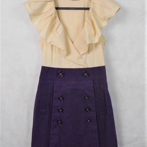 May include: A sleeveless dress with a cream colored top and a purple skirt. The top has a ruffled neckline and the skirt has a double row of buttons.