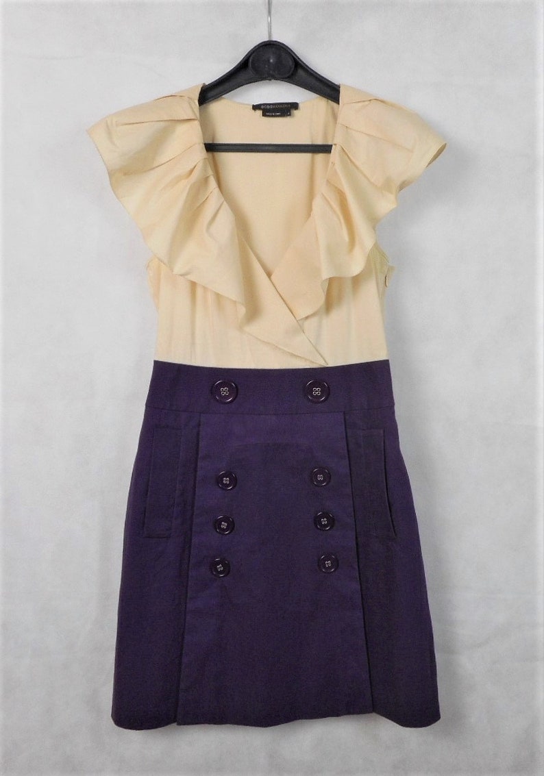 May include: A sleeveless dress with a cream colored top and a purple skirt. The top has a ruffled neckline and the skirt has a double row of buttons.