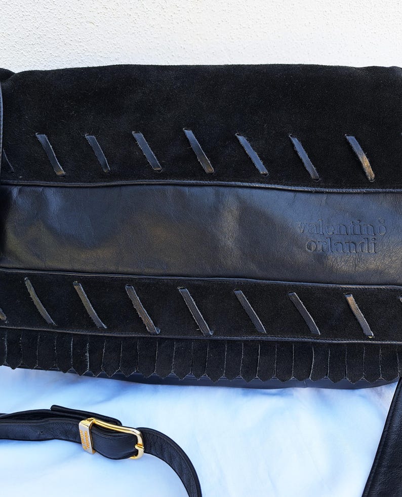 May include: A black handbag featuring a combination of smooth leather and suede textures. The front flap displays decorative leather lacing details and a fringe trim. The brand name valentino orlandi is embossed on the leather section. A black shoulder strap with a gold-tone metal buckle is attached.