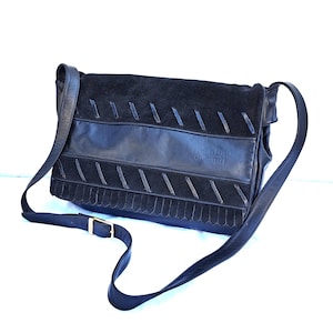 May include: A black leather and suede shoulder bag featuring a flap closure with diagonal stitched detailing, a fringe trim along the bottom edge, and an adjustable strap with a gold-tone metal buckle. The brand name Valentino Orlandi is embossed.