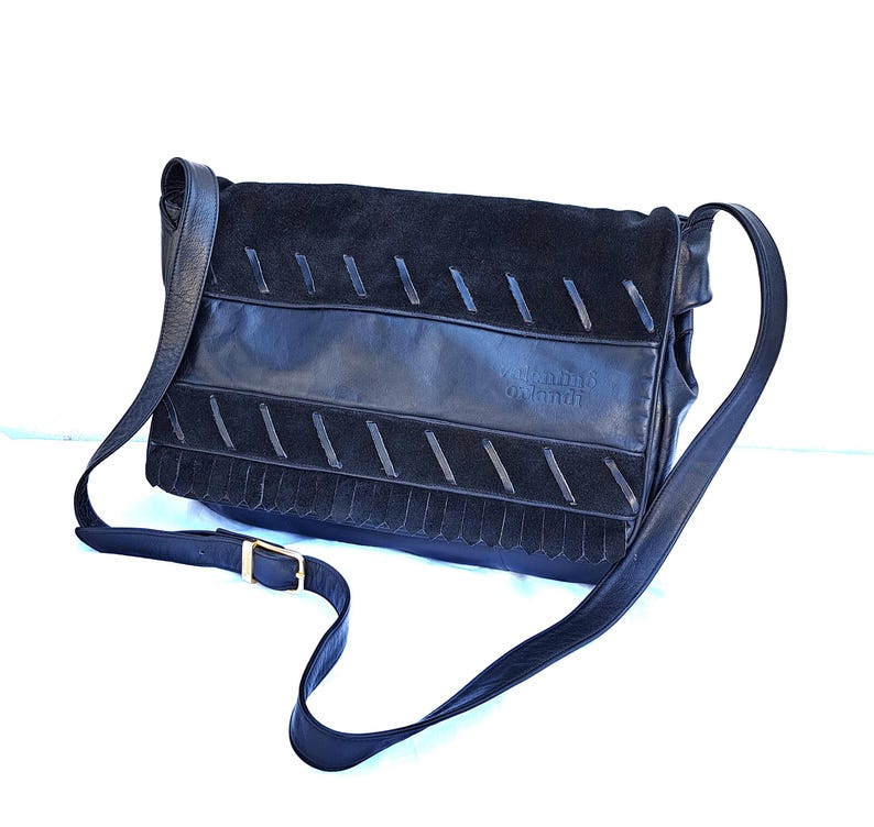 May include: A black leather and suede shoulder bag featuring a flap closure with diagonal stitched detailing, a fringe trim along the bottom edge, and an adjustable strap with a gold-tone metal buckle. The brand name Valentino Orlandi is embossed.