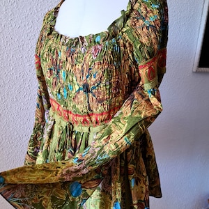 Könnte beinhalten: Eine grüne, braune und blaue Bluse mit floralem Muster, gerafftem Ausschnitt und langen Glockenärmeln. Die Bluse hat einen roten und grünen Besatz am Ausschnitt und an der Taille.