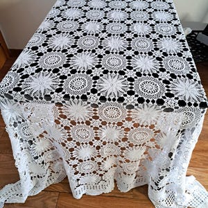 May include: White crocheted tablecloth with a delicate floral pattern. The tablecloth is draped over a table with a dark surface.