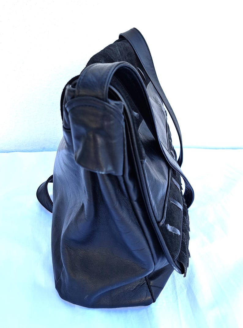 May include: A black leather shoulder bag featuring a fold-over flap with a textured suede panel and vertical stitched detailing. The bag includes a long, adjustable shoulder strap and a smooth leather body with a soft, slouchy construction.