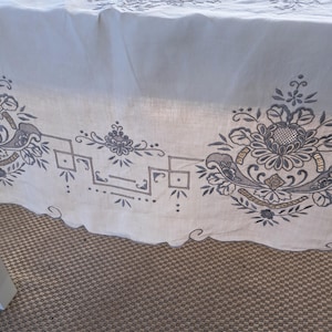 May include: White tablecloth with an embroidered border featuring a floral design in shades of gray and gold. The border includes geometric shapes and a scalloped edge.