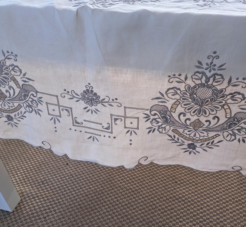 May include: White tablecloth with an embroidered border featuring a floral design in shades of gray and gold. The border includes geometric shapes and a scalloped edge.