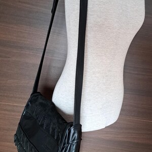 May include: A black shoulder bag featuring a flap with textured panels and decorative stitching. The bag has a long, adjustable black strap and a smooth finish on the lower section. The brand name is embossed on the front flap of the bag.