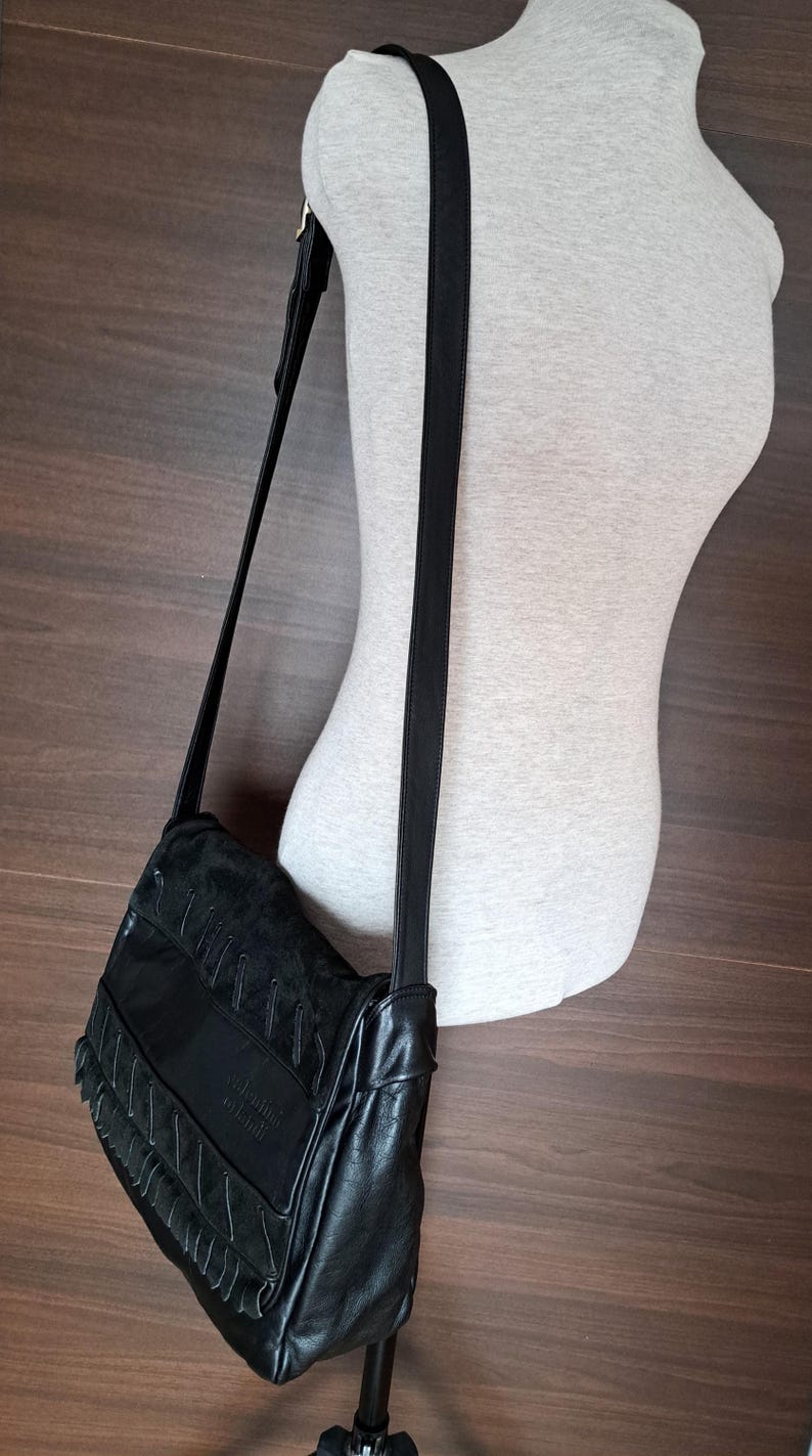 May include: A black shoulder bag featuring a flap with textured panels and decorative stitching. The bag has a long, adjustable black strap and a smooth finish on the lower section. The brand name is embossed on the front flap of the bag.