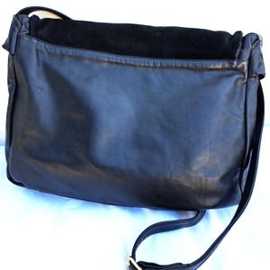 May include: A black leather messenger bag featuring a smooth finish on the main body and a textured suede flap closure. The bag includes a long, adjustable shoulder strap with a gold-toned metal buckle, providing a functional design.