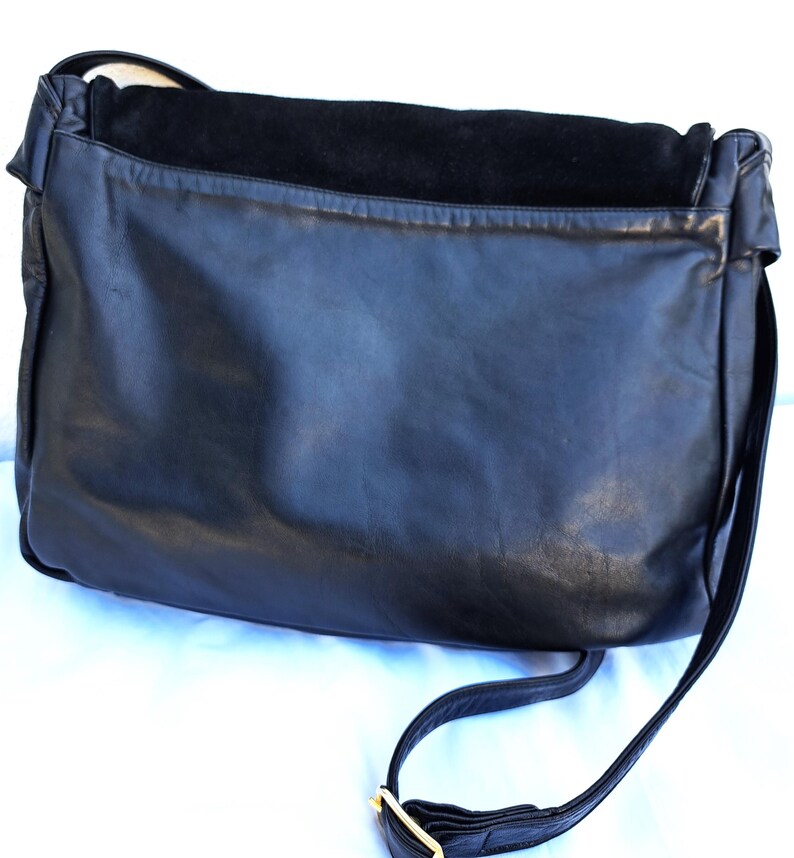 May include: A black leather messenger bag featuring a smooth finish on the main body and a textured suede flap closure. The bag includes a long, adjustable shoulder strap with a gold-toned metal buckle, providing a functional design.
