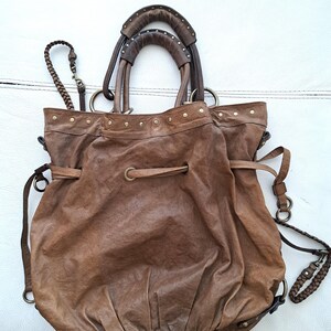 May include: A brown leather hobo bag featuring a drawstring closure, two rounded top handles with metal stud detailing, and a braided shoulder strap. The bag has a slouchy, gathered silhouette with brass-toned hardware and metal grommets.