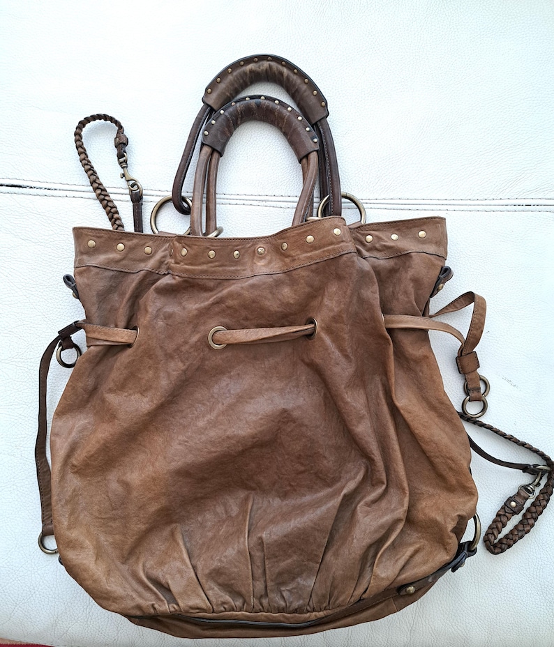 May include: A brown leather hobo bag featuring a drawstring closure, two rounded top handles with metal stud detailing, and a braided shoulder strap. The bag has a slouchy, gathered silhouette with brass-toned hardware and metal grommets.