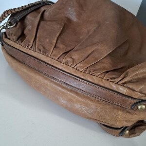 May include: A brown leather shoulder bag featuring a soft, gathered texture on the main body, a smooth leather trim along the base, and a braided leather strap attached with metallic hardware. The bag includes small brass-toned studs on the side.