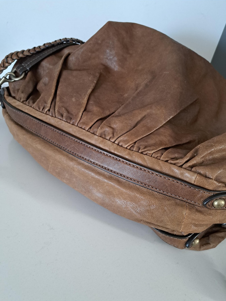 May include: A brown leather shoulder bag featuring a soft, gathered texture on the main body, a smooth leather trim along the base, and a braided leather strap attached with metallic hardware. The bag includes small brass-toned studs on the side.