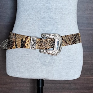 Könnte beinhalten: Ein Gürtel mit Schlangenmuster und einer silberfarbenen Schnalle und Akzenten. Der Gürtel ist um eine Schaufensterpuppe gewickelt, um sein Design zu präsentieren. Die Schnalle hat ein verziertes, graviertes Design. Das Muster des Gürtels weist Braun- und Beigetöne auf.