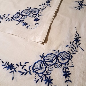 May include: White linen tablecloth with blue embroidered floral designs. The tablecloth features a repeating pattern of stylized flowers and leaves.