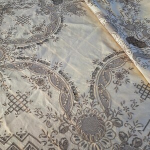May include: A white tablecloth with intricate embroidered floral patterns in a light brown color. The tablecloth has a delicate, vintage aesthetic.