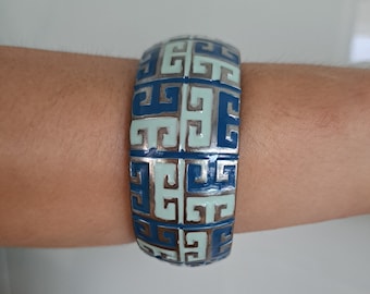 Può includere: Un braccialetto rigido in argento con un motivo geometrico blu e bianco. Il motivo è un disegno ripetitivo di quadrati intrecciati.