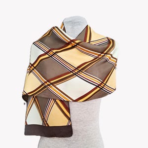 May include: A patterned scarf in shades of brown, cream, and yellow, draped over a mannequin. The scarf features a plaid design with intersecting lines of varying colours. The edges are finished with a solid brown border, creating a classic accessory.