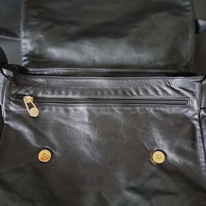 May include: A black leather bag featuring a fold-over flap with two gold-toned magnetic snap closures. The interior compartment includes a horizontal zippered pocket with a gold-toned zipper pull, providing secure storage for small items.