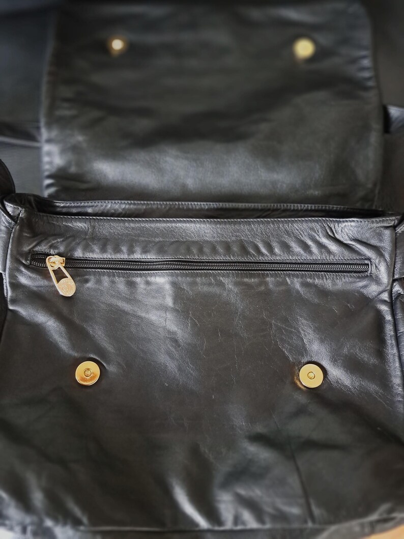 May include: A black leather bag featuring a fold-over flap with two gold-toned magnetic snap closures. The interior compartment includes a horizontal zippered pocket with a gold-toned zipper pull, providing secure storage for small items.
