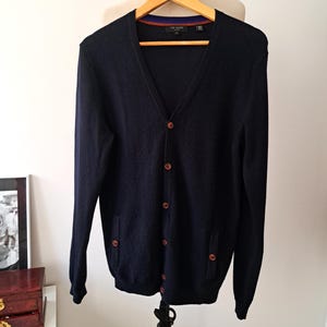 May include: A navy blue cardigan with a v-neck and button closure. The cardigan has two pockets and a brown trim on the inside of the collar.