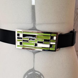 May include: A black leather belt with a silver buckle featuring a geometric design in green, white, and black.
