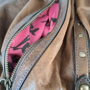 May include: A brown leather handbag featuring a zipped side pocket with a bright pink interior lining decorated with a black bird pattern. The exterior includes a vertical leather strip accented with four round brass-toned metal studs.