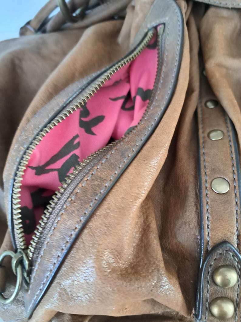 May include: A brown leather handbag featuring a zipped side pocket with a bright pink interior lining decorated with a black bird pattern. The exterior includes a vertical leather strip accented with four round brass-toned metal studs.