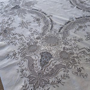 May include: A white tablecloth with intricate embroidered floral designs in a gray color. The embroidery features delicate leaves, flowers, and swirls.