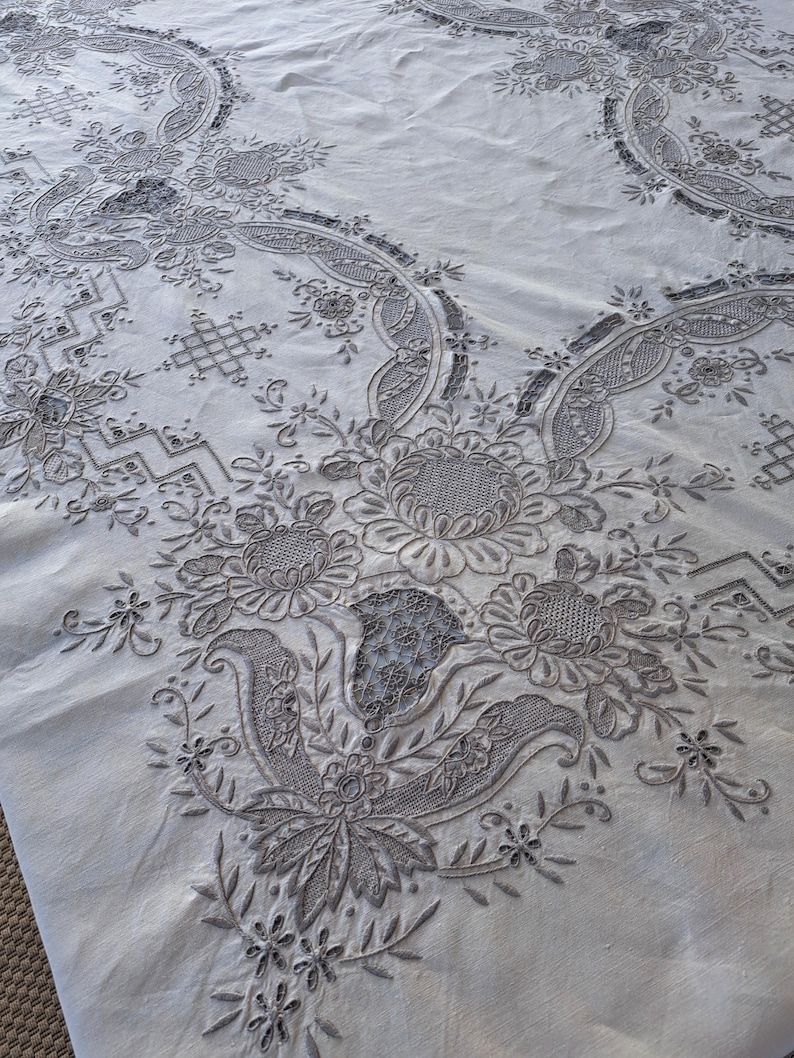 May include: A white tablecloth with intricate embroidered floral designs in a gray color. The embroidery features delicate leaves, flowers, and swirls.